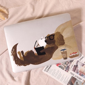May include: A white laptop cover featuring a brown dog wearing black glasses, lying down while reading a book. The dog is positioned next to a cup labeled MILK TEA and a small pastry. The Apple logo is integrated into the illustration as a book.