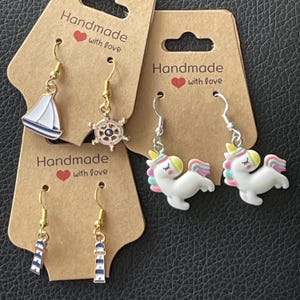 Puede incluir: Pendientes hechos a mano con temas náuticos y de unicornios. Los pendientes náuticos presentan un velero, un timón y un faro. Los pendientes de unicornio son blancos con crines y cuernos coloridos. Cada juego de pendientes está unido a una tarjeta marrón.