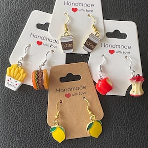 Könnte beinhalten: Eine Sammlung handgefertigter Modeschmuck-Ohrringe. Designs umfassen eine Pommes-Schachtel, einen Hamburger, Kaffeetassen, einen roten Apfel, einen Apfelkern und Zitronen. Jeder Ohrring ist an einer weißen oder braunen Karte mit dem Text "Handmade with love" befestigt.