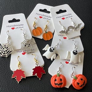 Peut inclure: Cinq paires de boucles d'oreilles pendantes sur le thème d'Halloween sur des cartes de présentation blanches marquées Handmade with love. Les motifs incluent des citrouilles orange, des toiles d'araignée, des chauves-souris, des fantômes et des feuilles.