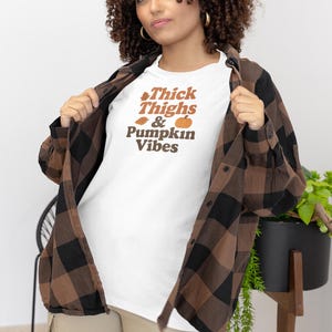 以下が含まれることがあります： 茶色とオレンジ色の「Thick Thighs & Pumpkin Vibes」の文字が入った白いTシャツ。茶色と黒のチェック柄フランネルシャツの下に着ています。モデルはカーキ色のパンツを着用しています。