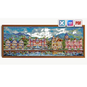 May include: A framed cross-stitch landscape depicting a colorful town by a lake, with mountains in the background. The buildings are in various colors, including red, yellow, and blue. The sky is a mix of blue and white. The frame is brown.