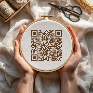 May include: A brown QR code cross-stitch design in a wooden embroidery hoop. The design is on a white fabric. The hoop is held by hands. Scissors and spools of thread are in the background.