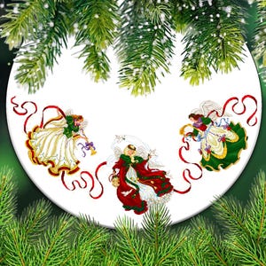 Puede incluir: Un plato decorativo blanco con tres ángeles con atuendos festivos. Cada ángel está representado con alas, vestidos fluidos y cintas rojas. El plato está rodeado de ramas de pino verdes, creando un tema navideño.