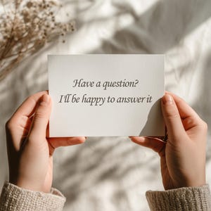 May include: A person holding a white card with the text "Have a question? I'll be happy to answer it" in black script. The card is held against a soft, neutral-toned background with dried flowers.
