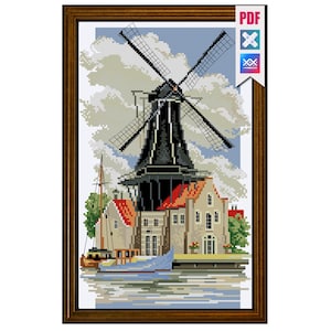 Puede incluir: Imagen de punto de cruz de un molino de viento holandés. El molino es negro con grandes aspas, sobre un cielo nublado. Los edificios con tejados rojos y un barco en el agua completan la escena, todo dentro de un marco de madera marrón.
