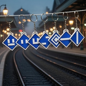 Peut inclure: Sept porte-clés avec des pendentifs en forme de losange bleu et blanc. Chaque pendentif présente un graphisme blanc différent sur fond bleu, attaché à une chaîne et un porte-clés argentés. Les porte-clés sont suspendus devant un arrière-plan flou de gare.