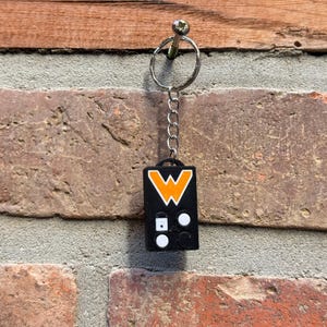 Op de afbeelding: Een zwarte sleutelhanger met een zilveren ketting en sleutelring. De sleutelhanger heeft een rechthoekige vorm met een oranje en wit "W" logo op de voorkant. Onder het logo staan twee witte cirkels en een witte vierkant.