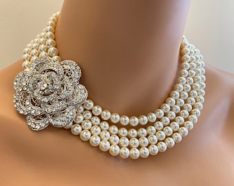 May include: A multi-strand necklace featuring three rows of cream-colored round pearls. A large, silver-toned floral brooch encrusted with clear, sparkling rhinestones is attached to the side, serving as a prominent focal point on the pearl strands.