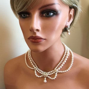 May include: A three-strand pearl necklace with a crystal and pearl pendant. The necklace is white and has a delicate design. The necklace is paired with matching pearl and crystal earrings.