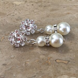 A pair of silver dangle earrings with a pearl and rhinestone design. Each earring features a cluster of small rhinestones in a flower shape, followed by a small pearl and a larger pearl with a row of rhinestones around the base.