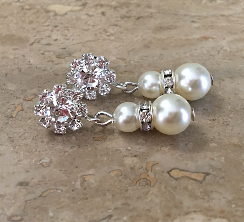 A pair of silver dangle earrings with a pearl and rhinestone design. Each earring features a cluster of small rhinestones in a flower shape, followed by a small pearl and a larger pearl with a row of rhinestones around the base.