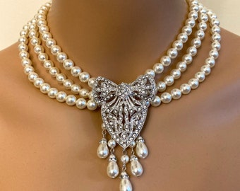 May include: A three-strand pearl choker necklace with a large, ornate, silver-toned pendant featuring a bow design and crystal embellishments. The pendant has a dangling drop of pearls.
