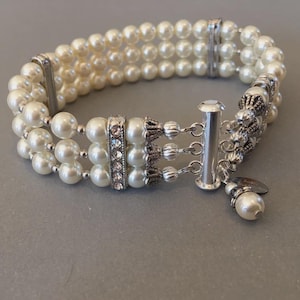 May include: A three-strand bracelet featuring cream-colored faux pearls, silver-toned metal spacers, and a central bar encrusted with clear rhinestones. The piece includes a silver-toned multi-strand clasp and a small dangling pearl charm.