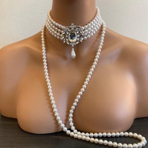 May include: A multi-strand white pearl choker necklace featuring a central oval rhinestone brooch with a dangling pearl drop, accompanied by a long, single-strand white pearl necklace that drapes down from the choker in a continuous loop.