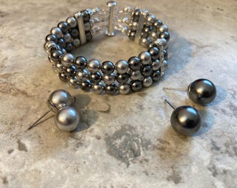 May include: A three-strand bracelet featuring alternating dark gray and light silver spherical beads, connected by silver-toned metal spacers and a cylindrical clasp. Two pairs of matching stud earrings with spherical beads are included.