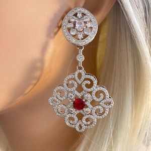 May include: A silver-toned drop earring featuring a circular top stud encrusted with clear stones. A large, square-shaped filigree pendant hangs below, decorated with clear stones and a single, vibrant round red stone at its center.