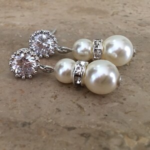 May include: A pair of silver earrings with a large clear rhinestone and two white pearl drops. The earrings have a silver chain connecting the rhinestone to the pearls.