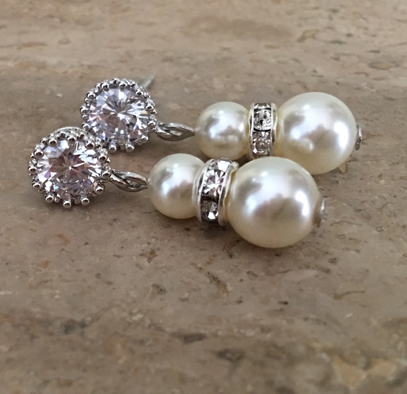 May include: A pair of silver earrings with a large clear rhinestone and two white pearl drops. The earrings have a silver chain connecting the rhinestone to the pearls.