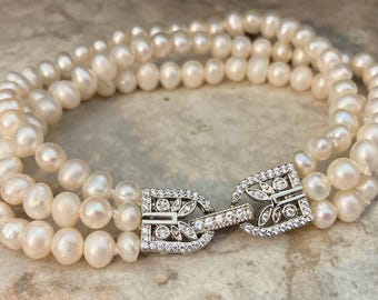 May include: A multi-strand bracelet composed of three rows of small, off-white, round freshwater pearls. The bracelet features a silver-toned metal clasp shaped like a stylized buckle, encrusted with small, clear, sparkling cubic zirconia stones.