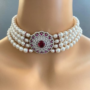 May include: A multi-strand pearl choker necklace featuring a central circular silver-tone clasp adorned with a large oval red gemstone, smaller red accent stones, and clear crystals, with red spacers between the white pearl strands.