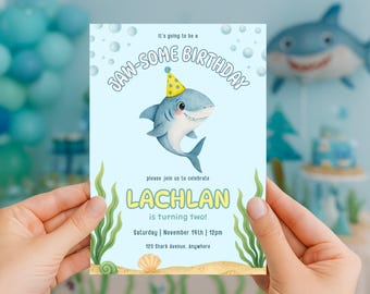 May include: A light blue birthday invitation with a cartoon shark wearing a party hat. The text says "JAW-SOME BIRTHDAY" and "LACHLAN is turning two!" with event details. The invitation is held by a person.