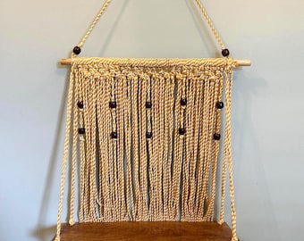 May include: A macrame wall shelf with a wooden shelf and decorative beads. The shelf is suspended by woven rope and features a natural beige colour. The shelf is designed for displaying small items and adds a bohemian touch to any room.
