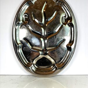May include: Oval silver-toned serving tray with a tree-like design in the center. The tray has a decorative beaded edge and several compartments. The surface reflects light, creating highlights and shadows.