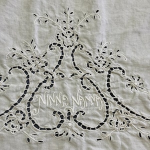 May include: Close-up of a white linen textile with intricate cutwork embroidery. The design features floral and scroll motifs, with the name "Ninna Ninna" embroidered in the center. The cutwork creates a delicate, open pattern.