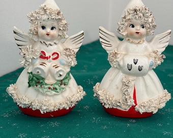 May include: Two ceramic angel figurines. Each angel has white wings, a white dress with a red base, and a decorative headpiece. One angel holds bells and holly, the other holds a decorated pouch. The figurines are vintage and festive.