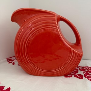 May include: A vibrant red ceramic pitcher with a rounded body and a curved handle. The pitcher features concentric, ridged detailing around its body. The spout is designed for easy pouring. The pitcher is a classic piece of kitchenware.