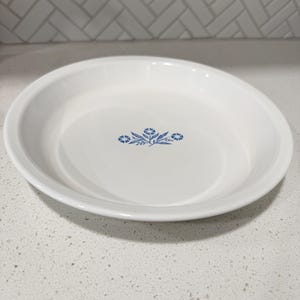 May include: A white ceramic pie dish with a shallow, circular design and a wide rim. The center of the dish features a small, blue floral pattern consisting of three stylized flowers with leaves, centered on the smooth, glossy white surface.