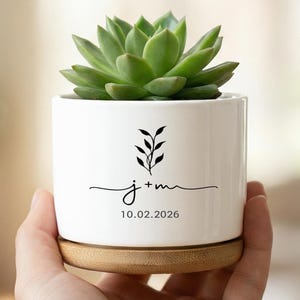 Può includere: Un vaso di ceramica bianco con una pianta succulenta verde. Il vaso ha una base in bambù e presenta un disegno floreale nero, le iniziali "j+m" e la data "10.02.2026".