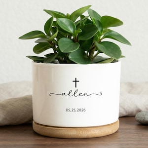 Könnte beinhalten: Ein weißer Keramik-Pflanztopf mit einem Kreuz, dem Namen "allen" und dem Datum 05.25.2026. Der Topf enthält eine grüne Blatpflanze und steht auf einem Holzsockel. Ein kleiner Terrakotta-Topf und graue Steine befinden sich im Hintergrund.