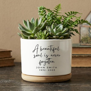 May include: White ceramic planter with a wooden base, filled with succulents and ferns. The planter has the inscription "A beautiful soul is never forgotten" with a name and dates. A decorative terrarium and books are in the background.