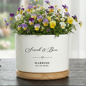 May include: A white ceramic planter with a wooden base, filled with colorful pansies and daisies. The planter is inscribed with "Sarah & Ben," a heart, and "MARRIED 05.10.2026."