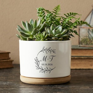 Könnte beinhalten: Ein weißer Keramik-Pflanztopf mit Holzsockel, gefüllt mit grünen Sukkulenten und Farnen. Der Topf hat ein schwarzes Blumengirlanden-Design mit den Initialen "M T" und dem Datum "10.18.2026". Der Topf steht auf einer Holzoberfläche.