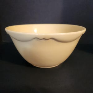May include: A cream-colored ceramic bowl with a scalloped rim. The bowl has a rounded shape and sits on a dark surface. This vintage-style bowl is suitable for serving or display.