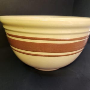 May include: Cream-colored ceramic mixing bowl with two wide, brown horizontal stripes. The bowl has a rounded shape and a slightly flared rim. A classic kitchenware item.