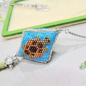May include: A beaded turtle charm with a blue background and orange shell, attached to a silver chain with a green tassel and a silver turtle charm.