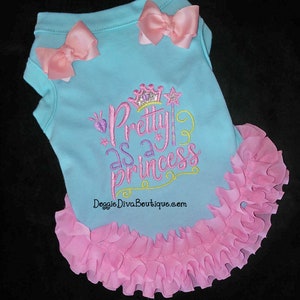 May include: A light blue dog shirt with pink bows and a ruffled hem. The shirt has the embroidered phrase "Pretty as a Princess" with a crown and star design. The website Doggie Diva Boutique.com is printed on the shirt.