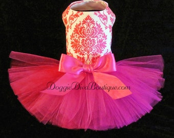 May include: A pink dog tutu with a white bodice featuring a red damask pattern. The tutu has a large pink bow and a layered tulle skirt. The text "Doggie Diva Boutique.com" is visible on the bow.
