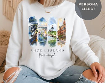 May include: White sweatshirt featuring watercolor illustrations of Rhode Island landmarks. The design includes images of the coast, sailboats, a lighthouse, and a building. The text "RHODE ISLAND Personalized" is printed below the images.