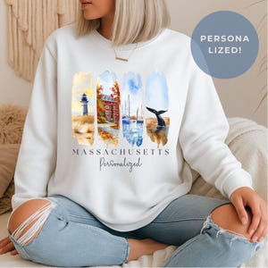 Könnte beinhalten: Weißes Sweatshirt mit einem Aquarelldesign mit einem Leuchtturm, einem Gebäude, Segelbooten und einer Walflosse. Das Wort "MASSACHUSETTS" ist unter dem Design aufgedruckt, darunter "Personalized". Ein blauer Kreis mit "PERSONALIZED!" befindet sich oben rechts.
