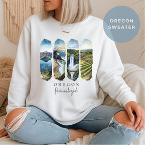 Peut inclure: Sweat-shirt blanc avec un motif aquarelle représentant des paysages de l'Oregon. Le motif comprend des images de montagnes, de cascades et de vignobles. Le mot "OREGON" est imprimé en dessous, avec "Personalized" en dessous. Un cercle bleu avec "OREGON SWEATER" est en haut à droite.