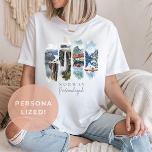 Op de afbeelding: Wit T-shirt met een aquarelontwerp van Noorse landschappen. Het ontwerp bevat bergen, watervallen en rode hutten. Het woord "NORWAY" staat boven "Personalized" gedrukt.