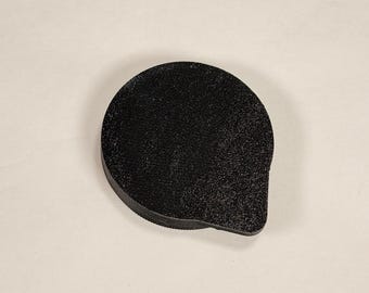 May include: A small, circular black object with a textured surface and a single protruding tab on one side. The item has a flat top and a rounded edge, appearing as a solid, singular piece with a matte finish and a consistent dark color throughout.