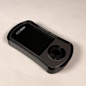 May include: A black and gray electronic device with a screen and buttons. The device has the word "COBB" printed on it. The device is rectangular with rounded edges and a circular button.