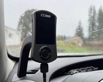 May include: A black COBB tuning device mounted on a car dashboard. The device has a screen and a central control button. A black cable connects the device to the car's system. The dashboard is dark gray.
