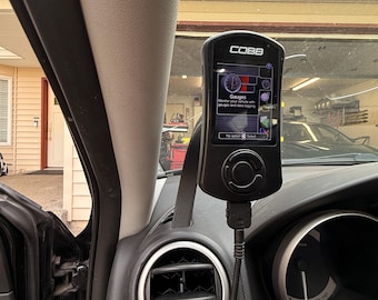 May include: A black Cobb Accessport automotive diagnostic device mounted on a vehicle dashboard. The screen displays a digital interface with the text Gauges, Monitor your vehicle with gauges and data logging, and No action Select, with a cable attached.